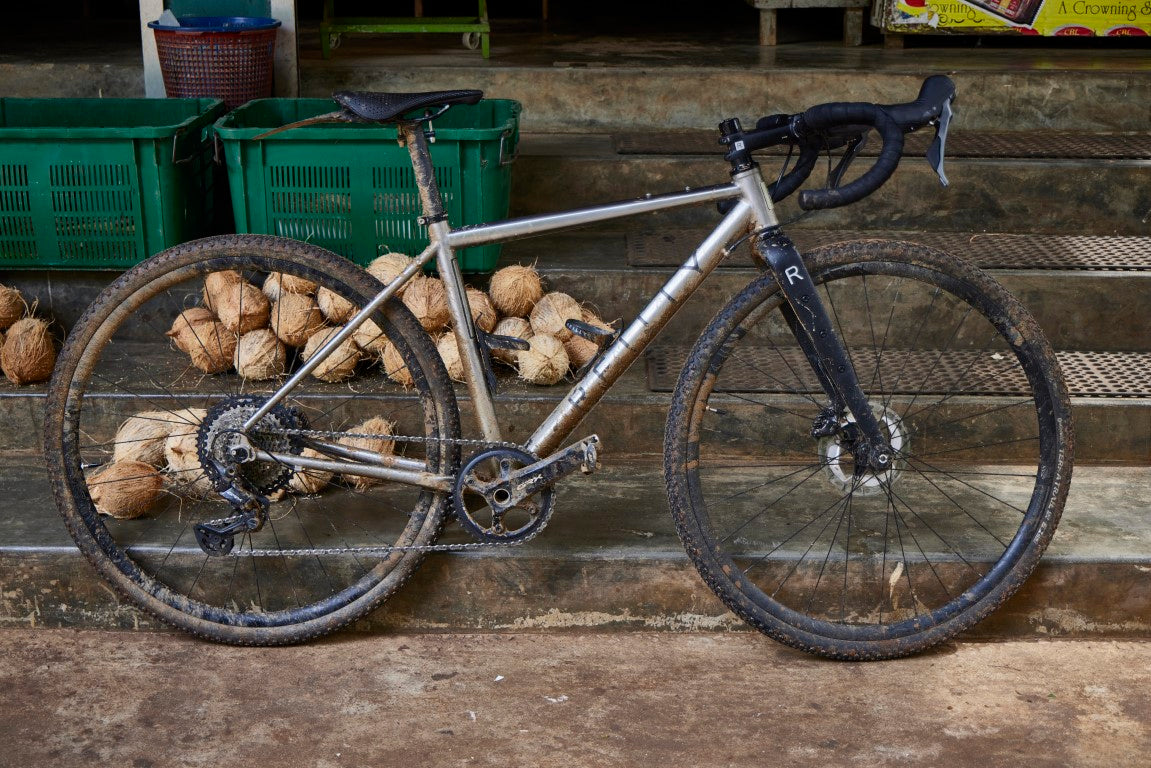 Extremly muddy, titanium gravel bike in front of steps with coconuts 