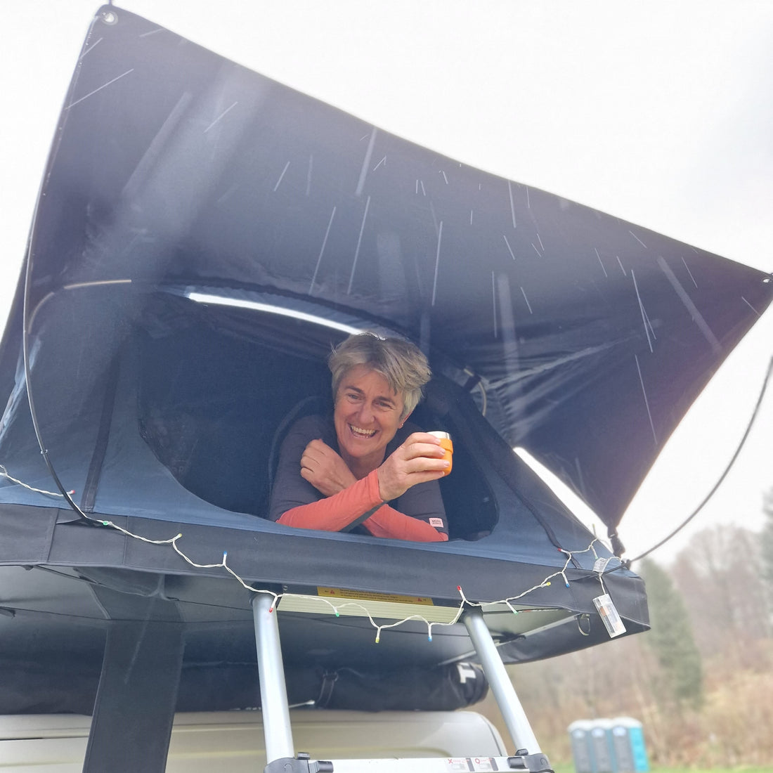 woman drinking coffee pokes her head out of the tent box ontop of a van.