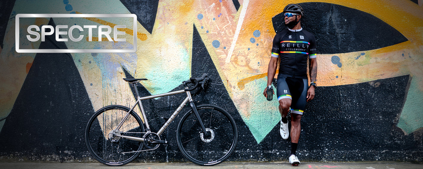 Man wearing dark glasses stants with foot up against a grafetti wall.  A gravel bike is resting against the wall next to him. SPECTRE  is top left.