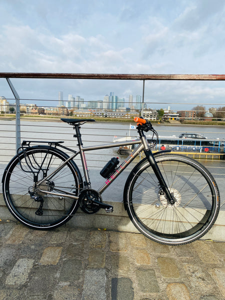 Gradient gravel bike with Pink logo and flat handlebars parked against a railing.  River Thames in the background with Canary Wharf in the distance  