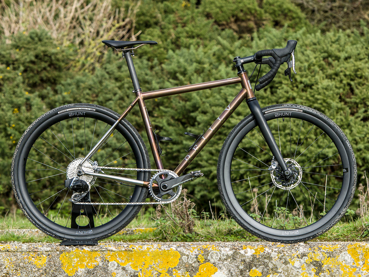 Gravel bike with brown anodised frame  on a wall with greenery as a background.