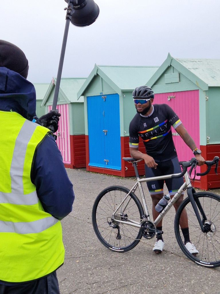 Man in Reilly kit leans on his titanium bike infront of beach huts.  A crew memeber in high vis jacket holding a light can be seen in the foreground.