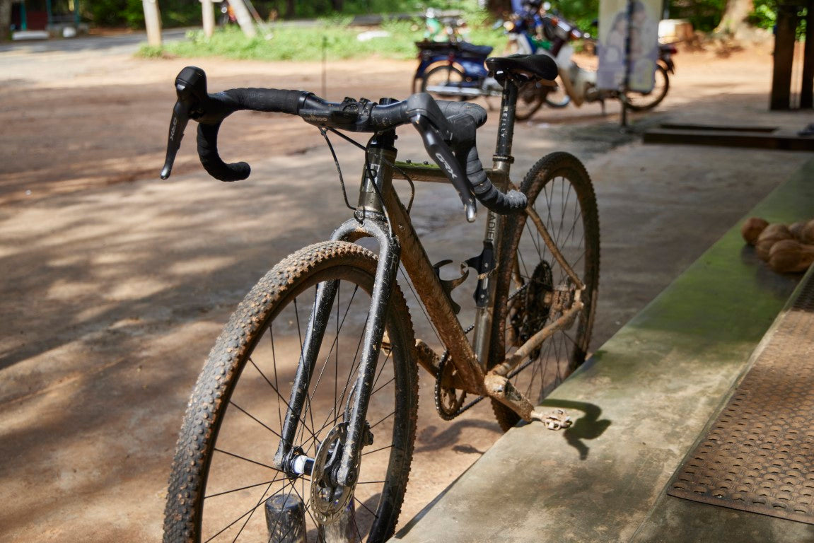 Front view of Reilly gravel bike, Gradient with muddy tyres parked against steps.  Coconuts can be seen on the steps and bare earth in the background.
