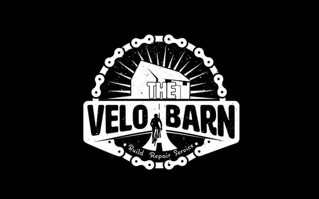 A bike chain with the image of a barn inside and a cyclist enroute to the barn.  The workding The Velo Barn is written across the logo.
