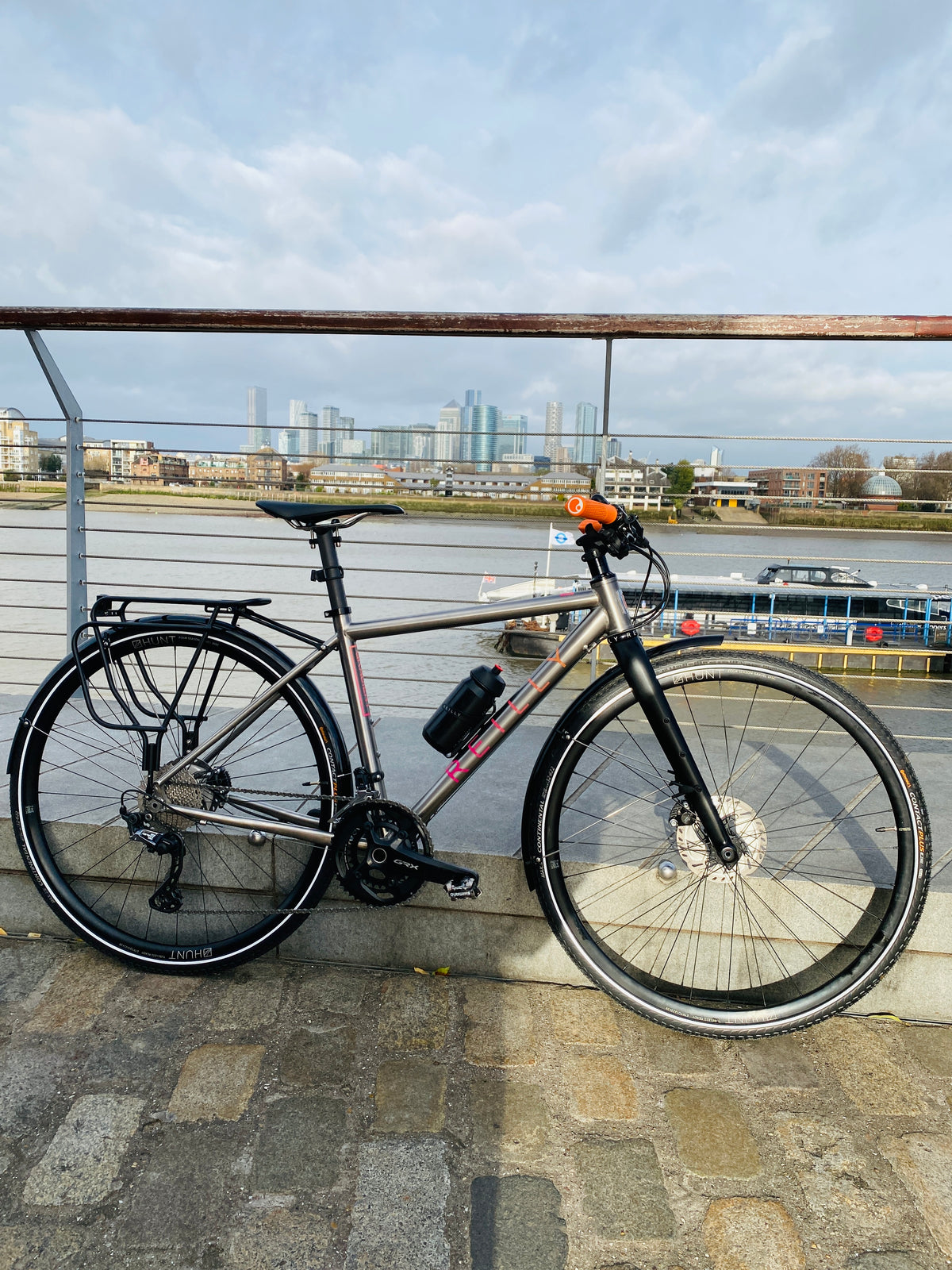 Gradient gravel bike with Pink logo and flat handlebars parked against a railing.  River Thames in the background with Canary Wharf in the distance  