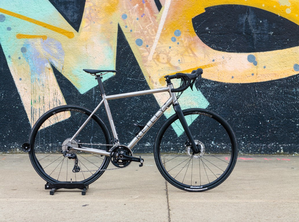 Titanium spectre against a wall with large yellow graffiti.  The bike  is brushed titanium with black logo and Strada wheels.